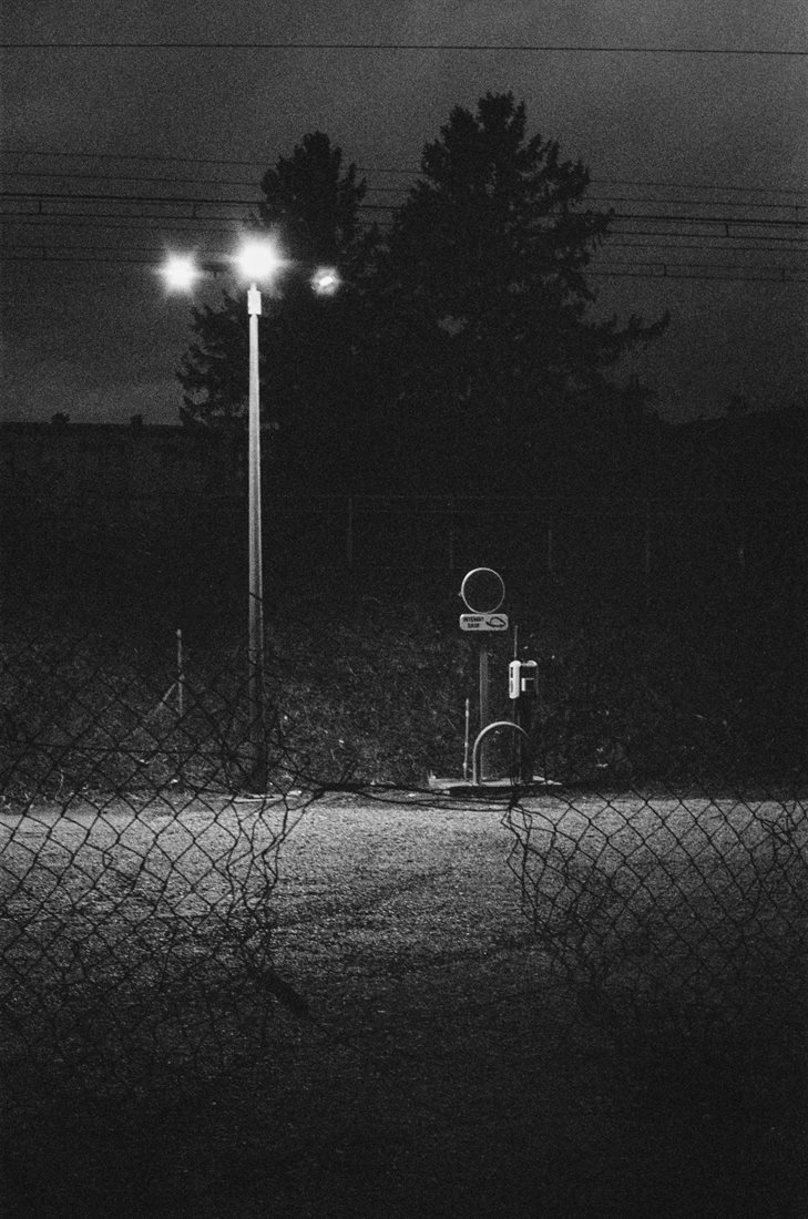 Montélimar, noir et blanc granuleux, photo de nuit, bord voie ferrée, nombreux fils électriques, partie centrale éclairée par trois projecteurs puissants perchés sur un haut poteau, parking vide, premier plan grillage simple torsion éventré